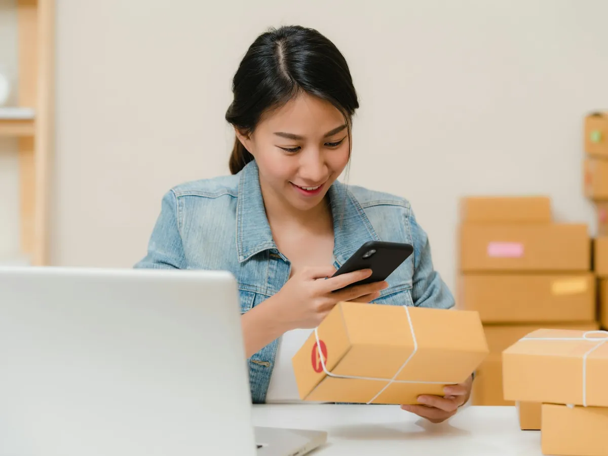 Mujer preparando un paquete para enviar mientras revisa su pedido en el celular